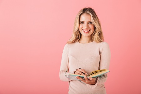 Portrait of a cheerful young woman wearing sweater standing isolated over pink background, writing notes in a diaryの写真素材