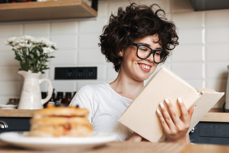 Smiling cheerful girl having tasty breakfast while sitting at the kitchen at home, reading a bookの写真素材