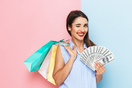 Cheerful young woman standing isolated over two colored background, holding money banknotes, carrying shopping bagsの写真素材