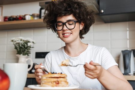Smiling cheerful girl having tasty breakfast while sitting at the kitchen at home, eating pancakesの写真素材