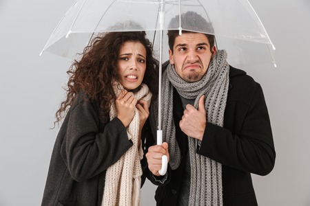 Upset frozen couple wearing autimn clothes standing under an umbrella isolated over gray backgroundの写真素材