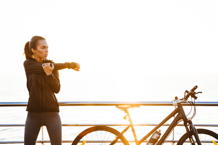Photo of sporty woman doing stretching exercises while standing near bicycle outdoors during sunrise...