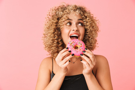 Photo of joyful curly woman 20s wearing dress eating donut while standing isolated over pink backgroundの写真素材