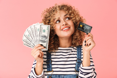 Photo of lovely curly woman 20s holding fan of dollar money and credit card while standing isolated over pink backgroundの写真素材
