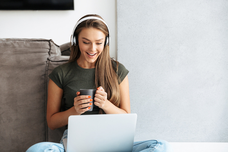 Cheerful young woman listening to music with headphones at home, working on laptop, drinking teaの写真素材