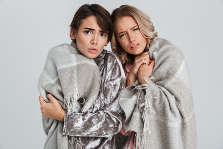 Two upset girls wearing pajamas standing isolated over gray background, wrapped in cozy blanketの写真素材