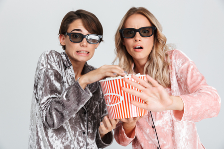 Two excited girls wearing pajamas standing isolated over gray background, watching a movie, eating popcornの写真素材