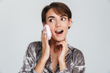 Pretty young girl wearing pajamas holding powder puff, isolated over gray backgroundの写真素材