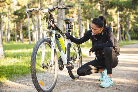 Attractive fit sportswoman having to fix her bicycle at the park, listening to music with wireless earphonesの写真素材