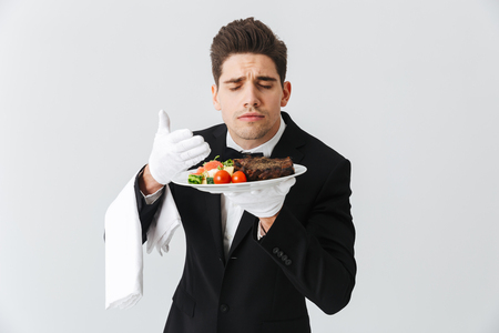 Portrait of a handsome young waiter in tuxedo showing beef steak dish on a plate over white backgroundの写真素材
