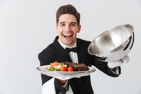 Portrait of a handsome young waiter in tuxedo showing beef steak dish on a plate over white...