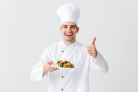 Excited man chef cook wearing uniform showing fresh green salad on a plate isolated over white background, thumbs upの写真素材