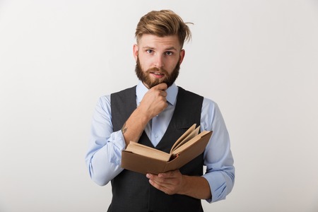 Image of a handsome young bearded man standing isolated over white wall background holding book...