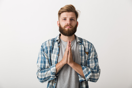 Worried bearded man dressed in plaid shirt standing isolated over white background, begging for somethingの写真素材