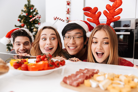 Group of cheerful friends celebrating Christmas at the kitchen at home, looking out from under the table at tasty foodの写真素材