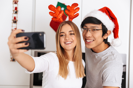 Image of young couple man and woman wearing christmas outfit standing in bright kitchen and taking selfie photo on mobile phoneの写真素材