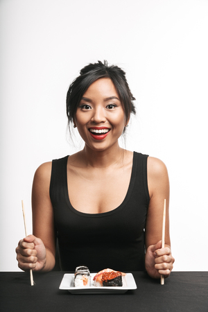 Pretty happy asian woman sitting at the table eating sushi from a plate isolated over white backgroundの写真素材