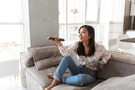 Photo of fashion asian woman 20s holding remote control and watching TV while sitting at sofa in cozy apartmentの写真素材