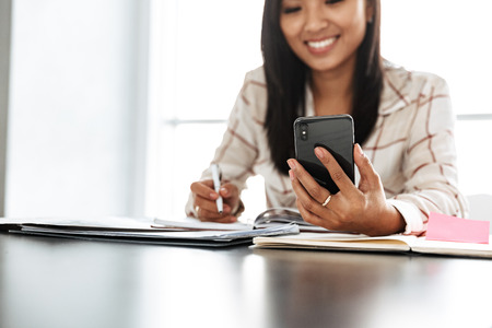 Image of attractive asian woman 20s using mobile phone while sitting at table indoor and working with documentsの写真素材
