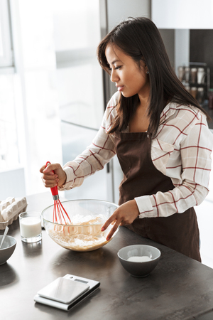 Photo of beautiful asian woman 20s smiling and making breakfast in bright kitchen interiorの写真素材