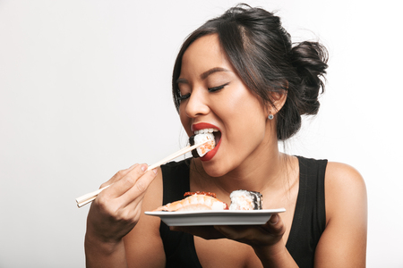 Pretty happy asian woman sitting at the table eating sushi from a plate isolated over white backgroundの写真素材