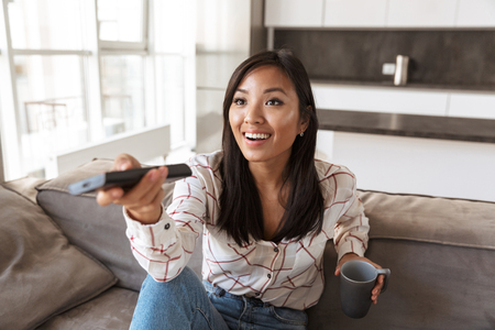 Photo of positive asian woman 20s holding remote control and watching TV while sitting at sofa in cozy apartmentの写真素材