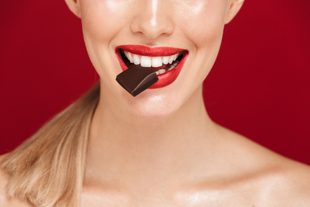 Cropped image of a pretty young topless woman standing isolated over red background, biting chocolateの写真素材
