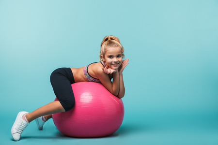 Cheerful little girl wearing sport clothes leaning on a fitness ball isolated over blue backgroundの写真素材