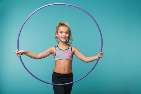 Cheerful little sports girl doing exercises with a hula hoop isolated over blue backgroundの写真素材