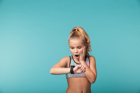 Excited little girl wearing sport clothing using smart watch isolated over blue background