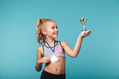 Cheerful little sports girl celebrating the win isolated over blue background, wearing a gold medal, showing a trophyの写真素材