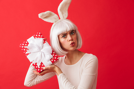 Cheerful young girl wearing Christmas bunny ears standing isolated over red background, holding gift boxの写真素材