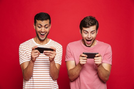 Two cheerful men friends standing isolated over red background, playing games on mobile phones