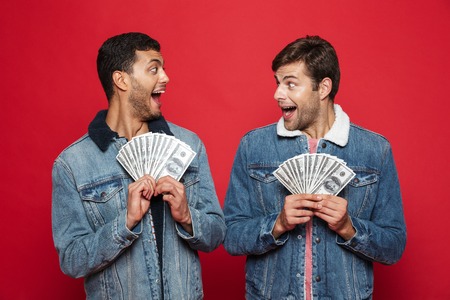 Two cheerful young men standing isolated over red background, holding money banknotesの写真素材