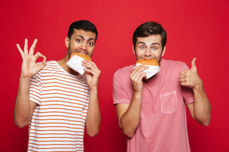 Two cheerful men friends standing isolated over red background, eating burgersの写真素材