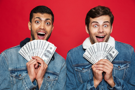 Two cheerful young men standing isolated over red background, holding money banknotesの写真素材