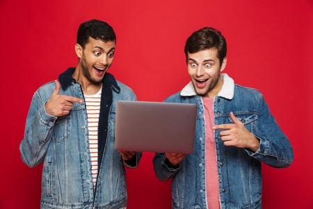 Two cheerful men friends standing isolated over red background, holding laptop computer, celebrating