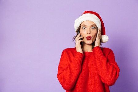 Portrait of an excited happy young woman wearing christmas hat isolated over purple background talking by mobile phone.の写真素材