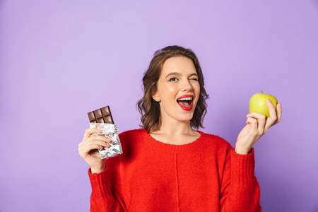 Image of a shocked emotional young woman posing isolated over purple background wall holding chocolate and apple.の写真素材