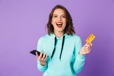 Image of a happy young woman posing isolated over purple background wall using mobile phone holding credit card.の写真素材
