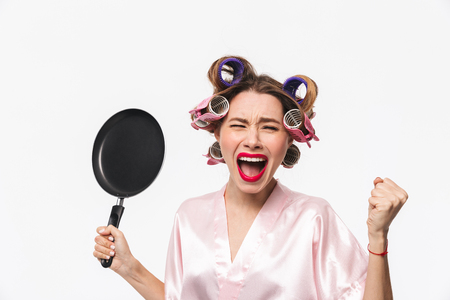 Angry housewife with curlers in hair standing isolated over white background, holding frying panの写真素材