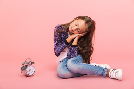 Pretty little girl sitting isolated over pink background, sleeping while sitting with an alarm clockの写真素材