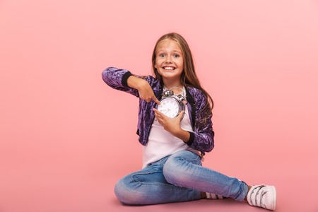 Pretty little girl sitting isolated over pink background, holding alarm clock, pointing finger