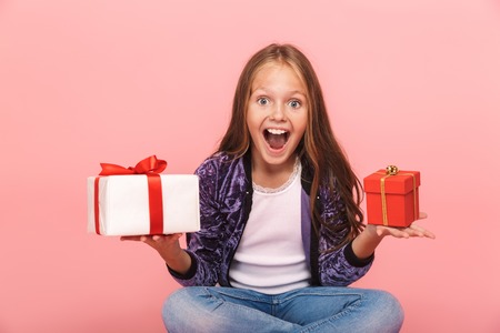 Excited pretty little girl sitting isolated over pink background, choosing between small and big gift boxesの写真素材