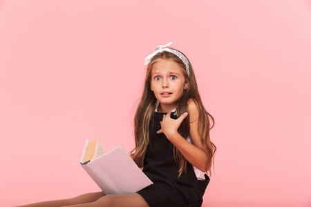 Surprised little girl wearing dress reading a book isolated over pink backgroundの写真素材