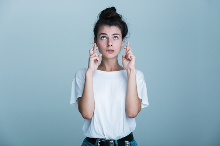 Portrait of a casual beautiful young girl isolated over blue background, holding fingers crossed for good luckの写真素材