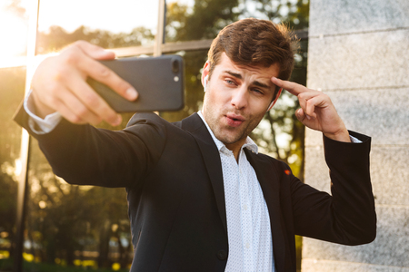 Photo of caucasian executive manager in suit holding mobile phone for selfie while standing outdoor against buildingの写真素材