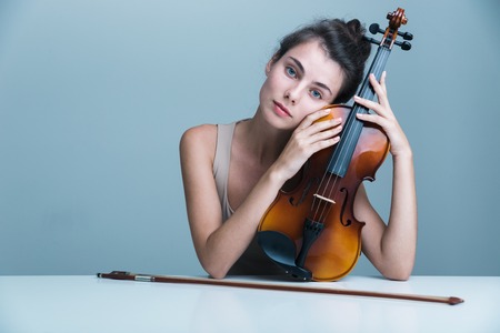 Portrait of a beautiful young woman sitting at the table with a violin isolated over blue backgroundの写真素材