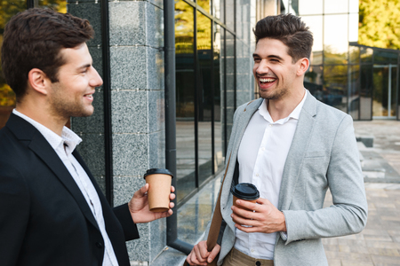 Photo of young entrepreneurs in suits standing outdoor near building with takeaway coffee during sunny dayの写真素材
