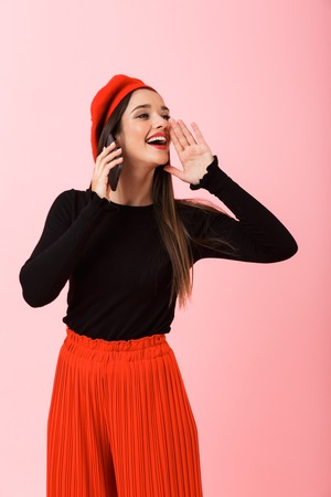 Portrait of a happy beautiful young woman wearing red beret standing isolated over pink background,...